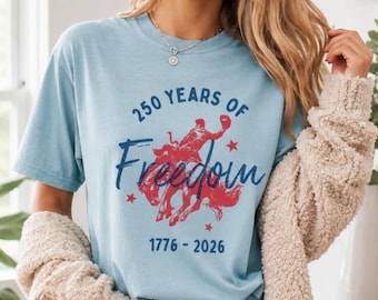 May include: Light blue t-shirt with a red and blue graphic celebrating 250 years of freedom. The design features a cowboy riding a horse, the words "250 Years of Freedom" and the years 1776-2026.