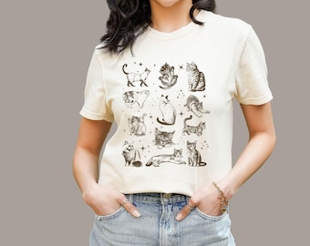 May include: A cream-colored short-sleeve t-shirt featuring a central graphic print of twelve distinct cats in various poses, rendered in a dark brown ink style. The design includes small star accents scattered among the feline illustrations.