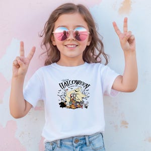 Op de afbeelding: Wit T-shirt met een Halloween-thema grafiek met de tekst "Welcome to Halloween". De afbeelding bevat personages en elementen die bij de feestdag horen. Het shirt wordt door een jong persoon gedragen.