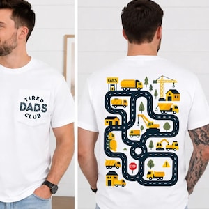 May include: White t-shirt with a pocket featuring the text "TIRED DADS CLUB" in navy blue. The back of the shirt has a colorful road map design with yellow trucks, houses, and a crane. A fun, casual tee.