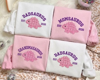 Könnte beinhalten: Vier langärmelige Sweatshirts in Weiß und Hellrosa mit violetter Stickerei und einer rosa Dinosauriergrafik. Die Designs enthalten die Wörter DADSAURUS, MOMSAURUS und GRANDMASAURUS, jeweils ergänzt durch den Text EST 2026.