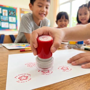May include: A red and gray self-inking rubber stamp with a clear base, being pressed onto a white sheet of paper. The stamp leaves a red circular impression featuring a smiling star icon and the text "Mrs. Amanda Says You are a star!"