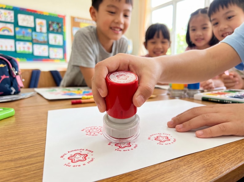 May include: A red and gray self-inking rubber stamp with a clear base, being pressed onto a white sheet of paper. The stamp leaves a red circular impression featuring a smiling star icon and the text "Mrs. Amanda Says You are a star!"