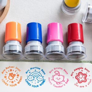 May include: Four self-inking stamps with orange, blue, pink, and red caps. Each stamp creates a unique circular impression on paper: Mrs. Rafael Says Loves Your Work, Mrs. Martina Says Loves Your Work with a rainbow, Mrs. Kristin Says Happy Teacher's Day with an apple, and Mrs. Amanda Says You Are A Star with a star.