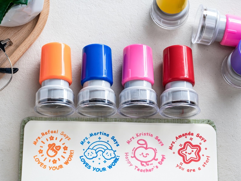 May include: Four self-inking stamps with orange, blue, pink, and red caps. Each stamp creates a unique circular impression on paper: Mrs. Rafael Says Loves Your Work, Mrs. Martina Says Loves Your Work with a rainbow, Mrs. Kristin Says Happy Teacher's Day with an apple, and Mrs. Amanda Says You Are A Star with a star.