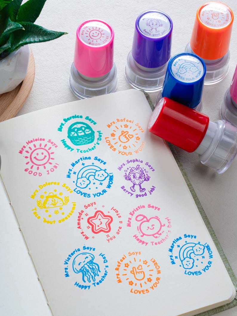 May include: A collection of colorful self-inking stamps with matching ink impressions on paper. The stamps feature various designs including a sun, rainbow, strawberry, taco, star, apple, and jellyfish, each with personalized teacher names and phrases.