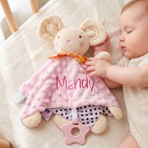 May include: A plush mouse security blanket featuring a soft pink dimpled fabric with the name Mandy embroidered in pink thread. The mouse has cream-colored limbs, a colorful floral neck ribbon, a black and white polka dot underside, and a star-shaped teether.