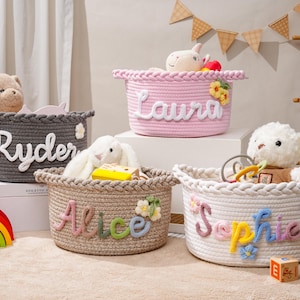 May include: Four round woven rope storage baskets featuring braided rims and personalized names in raised lettering. The baskets are colored dark gray, light pink, tan, and white, each decorated with small fabric flowers and filled with plush toys.
