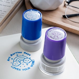May include: Two self-inking stamps with blue and purple plastic caps. The blue stamp features a rainbow and cloud design with the text 'Mrs. Martina Says LOVES YOUR WORK'. The purple stamp has a strawberry character and the text 'Mrs. Sophia Says Berry good job'.