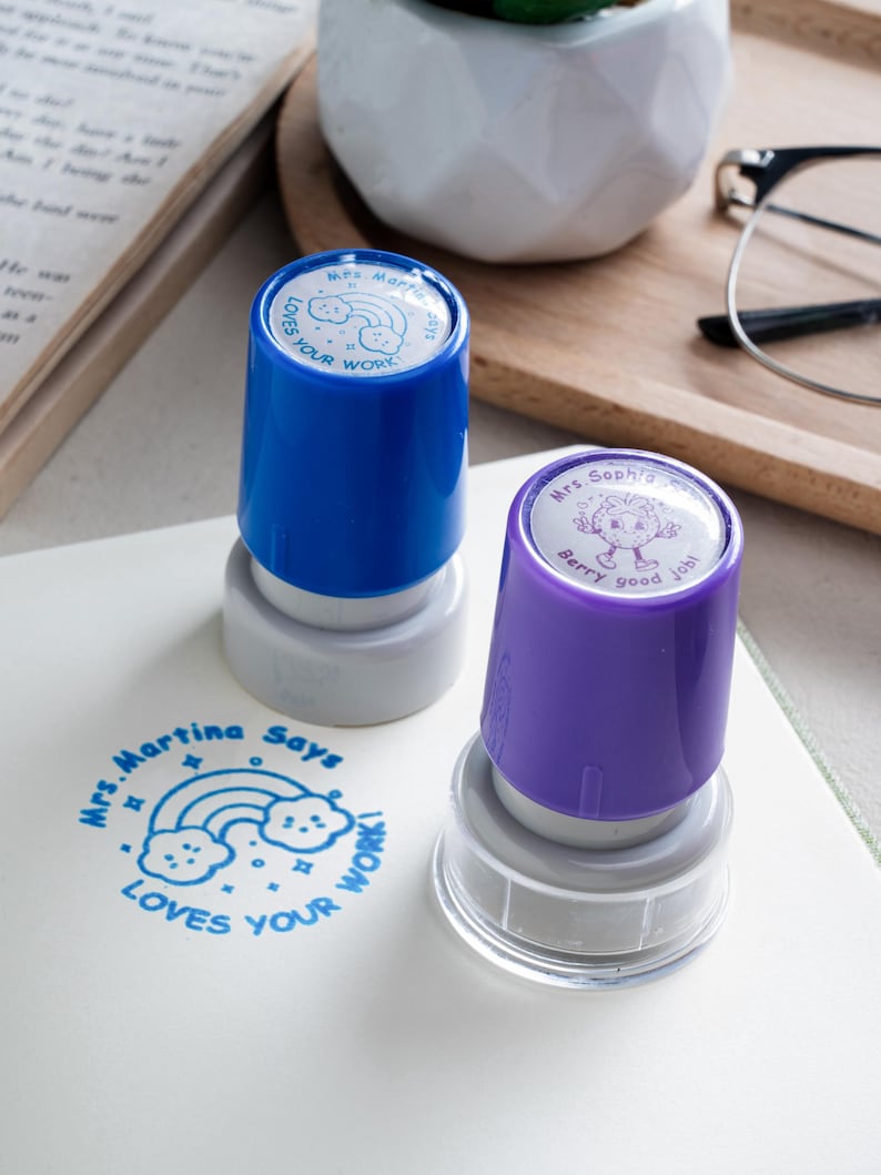 May include: Two self-inking stamps with blue and purple plastic caps. The blue stamp features a rainbow and cloud design with the text 'Mrs. Martina Says LOVES YOUR WORK'. The purple stamp has a strawberry character and the text 'Mrs. Sophia Says Berry good job'.