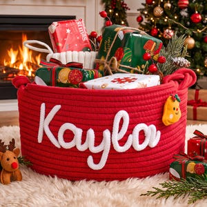 May include: A round red rope basket featuring the white knitted name Kayla on the front. The basket is filled with various wrapped Christmas gifts, including red and green packages, and is decorated with a small yellow gingerbread man ornament.