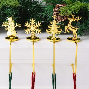 May include: Four gold-colored Christmas stocking holders with festive designs: a snowman, snowflake, Christmas tree, and reindeer. Each holder has a long, curved hook with a loop for hanging stockings. Green and red yarn hang from the hooks.