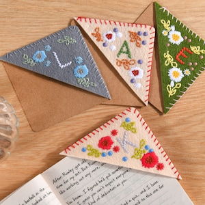 Peut inclure: Ensemble de marque-pages d'angle en feutre faits à la main avec des motifs floraux brodés. Les marque-pages sont de différentes couleurs, dont le gris, le beige et le vert, avec des coutures rouges, blanches et bleues. Un marque-page porte la lettre "A".