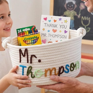 Puede incluir: Una cesta redonda de cuerda tejida en color blanco roto con el nombre Mr. Thompson bordado en hilo marrón, azul, verde y naranja. Dentro de la cesta hay una caja amarilla de 24 ceras Crayola y una tarjeta blanca con corazones y estrellas de colores que dice Thank You Mr. Thompson.