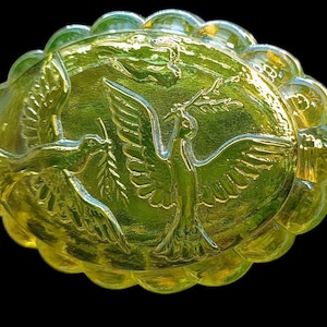 May include: A yellow-green glass dish with an oval shape and a scalloped edge. The dish features embossed images of doves carrying olive branches. The background is a darker green, and the dish is set against a black background.