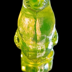 Op de afbeelding: Een doorschijnende, groene gummybeer snoep tegen een zwarte achtergrond. De gummybeer staat, met details van zijn vorm zichtbaar. Het snoepje heeft een glanzend uiterlijk, met licht dat weerkaatst op het oppervlak.