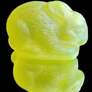 Op de afbeelding: Een limoengroene glazen konijnenfiguur met een glad, reflecterend oppervlak. Het konijn zit in een rusthouding, met de oren naar achteren. De achtergrond is zwart.