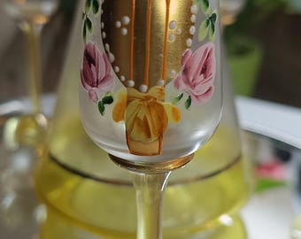 May include: A clear glass cordial glass with a gold rim and stem, decorated with pink roses and gold accents. The glass is part of a set, with a matching decanter visible in the background.