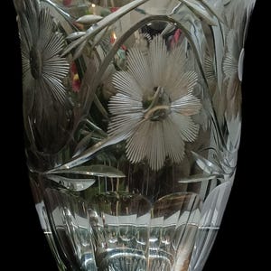 Può includere: Un vaso di vetro trasparente con disegni floreali incisi. Il vaso presenta un bordo ampio e smerlato e una base dettagliata. Il motivo floreale comprende grandi fiori stilizzati e foglie.