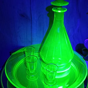 Può includere: Una caraffa di vetro verde brillante con tappo abbinato, posta su un vassoio rotondo. Tre piccoli bicchierini sono disposti sul vassoio. Il vetro ha un'estetica vintage.