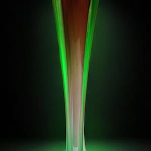 Puede incluir: Un florero de vidrio alto y esbelto con un exterior verde vibrante que transiciona hacia un interior rojo profundo. El florero tiene un borde acampanado de múltiples facetas y una base estructurada con cuatro patas, iluminado desde abajo.