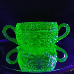Puede incluir: Un azucarero de vidrio verde con dos asas. El tazón tiene un diseño de vidrio cortado y está hecho de vidrio de uranio, que brilla bajo una luz negra.