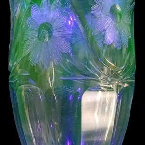 Könnte beinhalten: Eine klare Glasvase mit grün-blauer schillernder Oberfläche. Die Vase weist geätzte Blumendesigns auf, darunter große Blüten und Blätter. Die Vase hat einen breiten, gewellten Rand und einen abgerundeten Boden. Der Hintergrund ist schwarz.