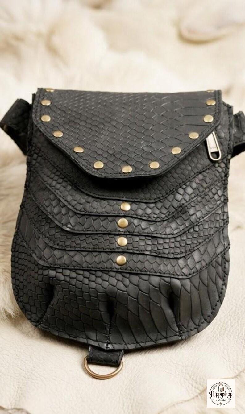 Black reptile texture leather harness bag with twin studded pockets and adjustable straps, photographed on faux fur background with vintage compass and journals.