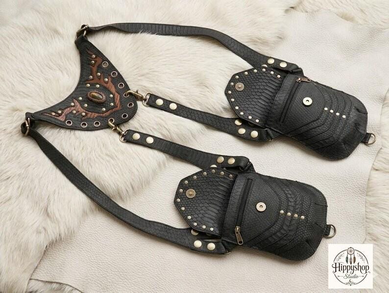 Alternative fashion leather chest bag with symmetrical studded pouches and textured finish, flat lay display with vintage compass, brown leather journals, coins, and flask.