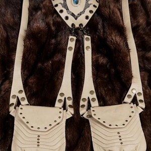 Statement leather festival harness bag with adjustable shoulder straps and studded pouch details, photographed on faux fur with travel compass, journals, and vintage bottle.