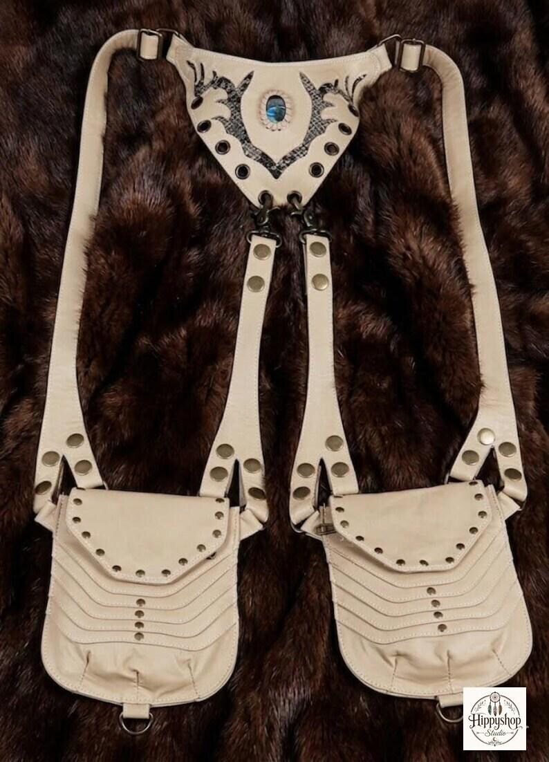 Statement leather festival harness bag with adjustable shoulder straps and studded pouch details, photographed on faux fur with travel compass, journals, and vintage bottle.