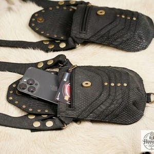 Handmade gothic leather harness bag with adjustable straps and studded detailing, arranged with vintage compass, brown journals, scattered coins, and corked bottle.