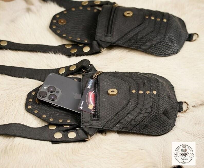 Handmade gothic leather harness bag with adjustable straps and studded detailing, arranged with vintage compass, brown journals, scattered coins, and corked bottle.