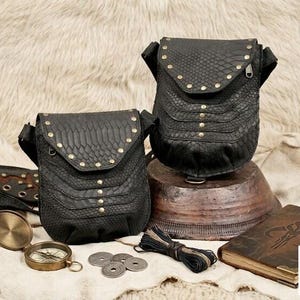 Festival ready black leather body harness bag with decorative studs and adjustable straps, paired with vintage travel accessories including compass, journals, coins, and flask.