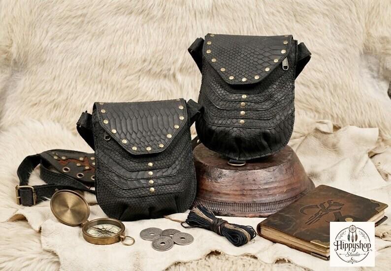 Festival ready black leather body harness bag with decorative studs and adjustable straps, paired with vintage travel accessories including compass, journals, coins, and flask.