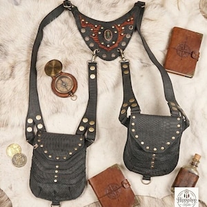 Black leather festival harness bag with two front pouches, brass studs, and adjustable straps, styled flat lay on faux fur with a compass, coins, leather journals, and a glass bottle.