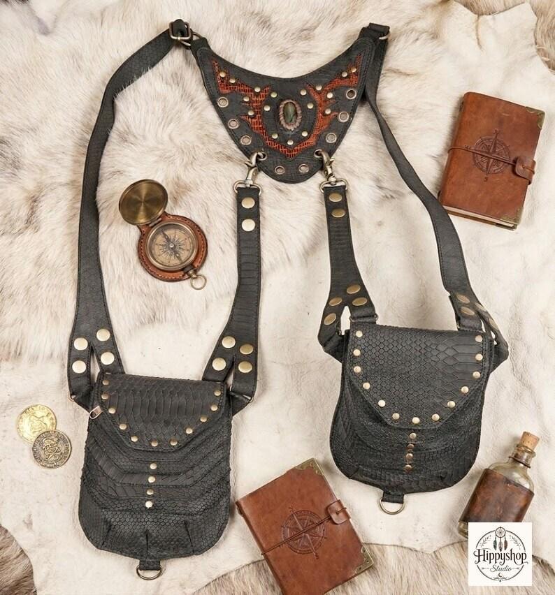 Black leather festival harness bag with two front pouches, brass studs, and adjustable straps, styled flat lay on faux fur with a compass, coins, leather journals, and a glass bottle.