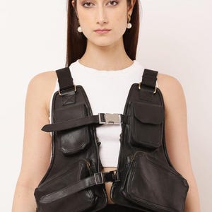 May include: A black utility vest featuring multiple storage pockets, adjustable straps, and metallic buckle closures. The garment is worn over a white sleeveless top and includes a small logo in the bottom right corner that reads Hippyshop Studio.