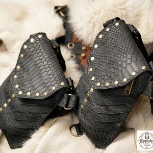 Gothic style leather chest harness bag with twin studded pouches and wide straps, arranged in flat lay with antique compass, embossed journals, coins, and corked glass flask.