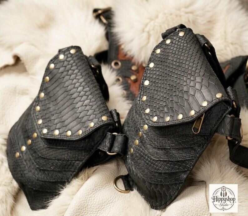 Gothic style leather chest harness bag with twin studded pouches and wide straps, arranged in flat lay with antique compass, embossed journals, coins, and corked glass flask.