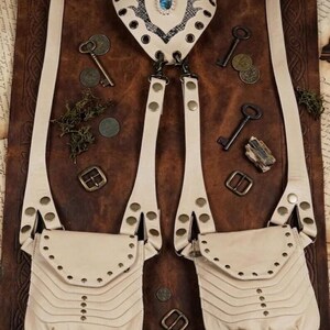 Festival fashion leather chest harness featuring dual front pouches and brass rivets, displayed on soft fur with embossed notebooks, compass, coins, and bottle.