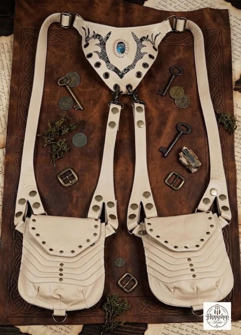 Festival fashion leather chest harness featuring dual front pouches and brass rivets, displayed on soft fur with embossed notebooks, compass, coins, and bottle.