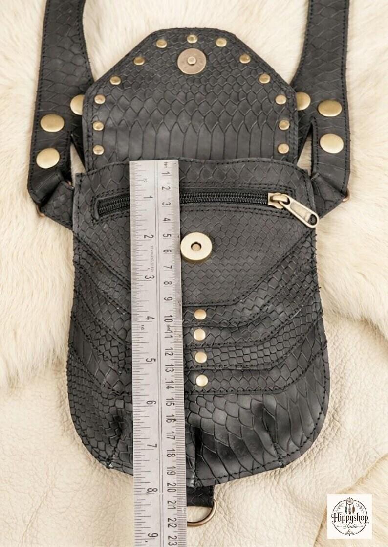 Handmade black leather utility harness featuring dual textured pouches and brass rivet detailing, displayed with vintage compass, brown journals, coins, and bottle on soft fur background.