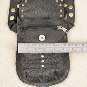 Explorer style leather body harness bag with decorative studded pouches and wide straps, flat lay with antique compass, embossed notebooks, coins, and glass flask