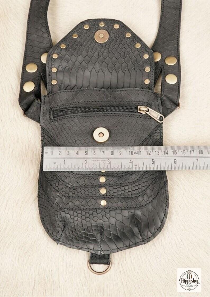Explorer style leather body harness bag with decorative studded pouches and wide straps, flat lay with antique compass, embossed notebooks, coins, and glass flask