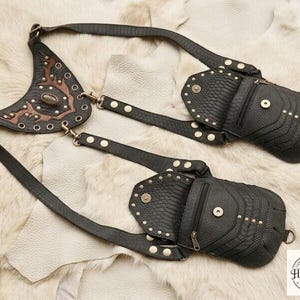 Rustic leather harness crossbody bag featuring two flap pouches and brass rivets, arranged with explorer themed props including compass, coins, journals, and cork bottle.