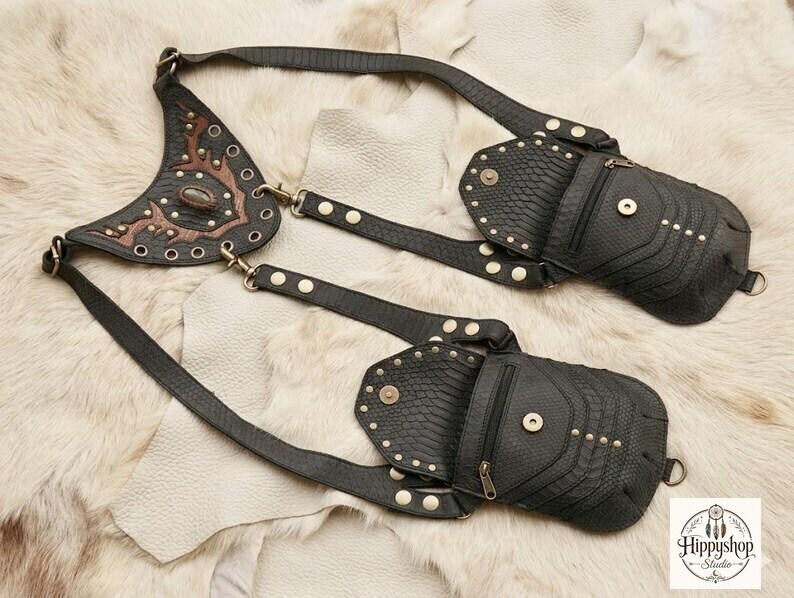 Rustic leather harness crossbody bag featuring two flap pouches and brass rivets, arranged with explorer themed props including compass, coins, journals, and cork bottle.