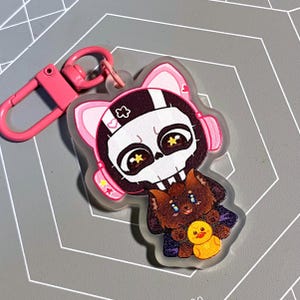 May include: A pink and white keychain featuring a cartoon skull character wearing headphones and holding a brown and white animal with a yellow duck.