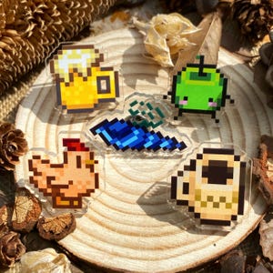 Peut inclure: Cinq épingles à thème pixel art représentant une chope de bière, un poulet, une créature verte, un objet bleu et blanc et une tasse à café. Les épingles sont sur une surface en bois.