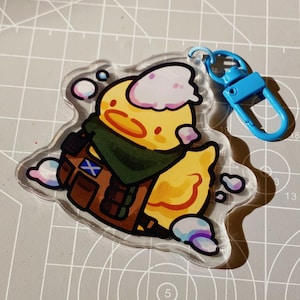 May include: A clear acrylic keychain featuring a yellow cartoon duck wearing a green scarf and brown backpack with a blue flag. The keychain has a blue clasp and is surrounded by white and purple bubbles.